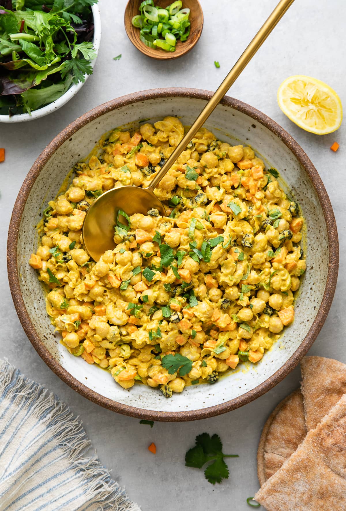 top down view of bowl with freshly made healthy curried chickpea salad with serving spoon with items surrounding.